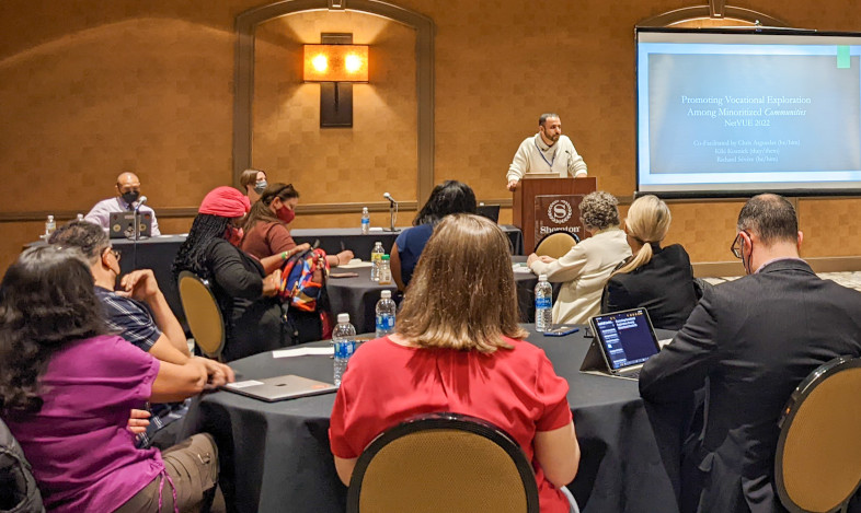 Presenter speaks from podium beside projector screen with seated participants