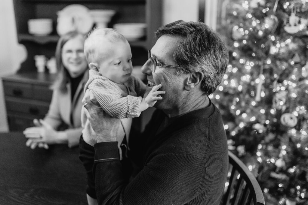 John Barton, smiling, holding up his infant grandson who reaches for his face.