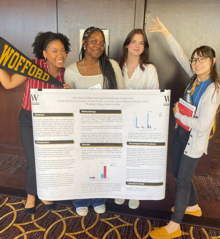 Four Wofford students standing with their research sign and a Wofford pennant