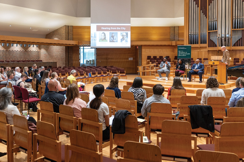 Panelists speak from stage in front of large organ pipes to seated participants