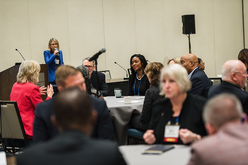 Participants discuss topics at roundtables with presenter standing at podium