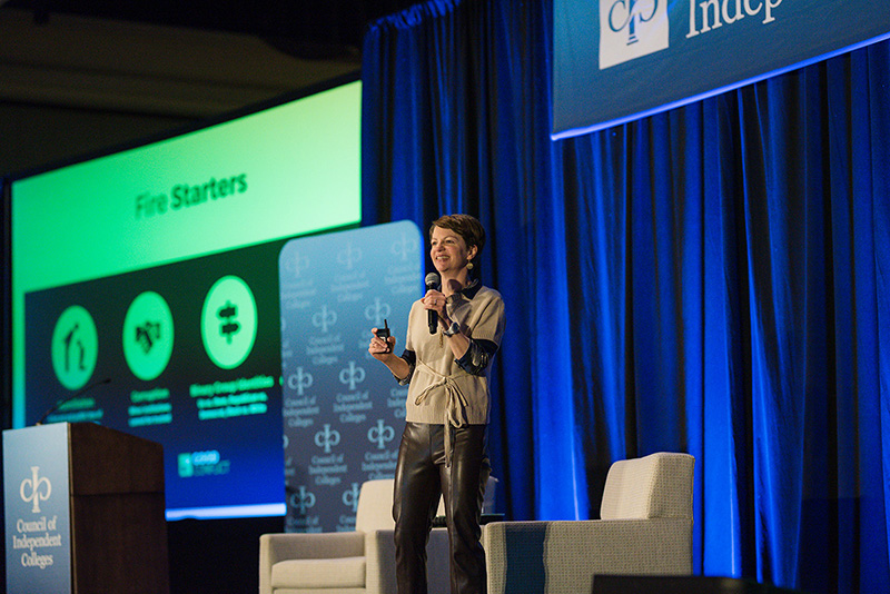 Amanda Ripley stands on stage in front of projection screen holding a microphone and slide advancer