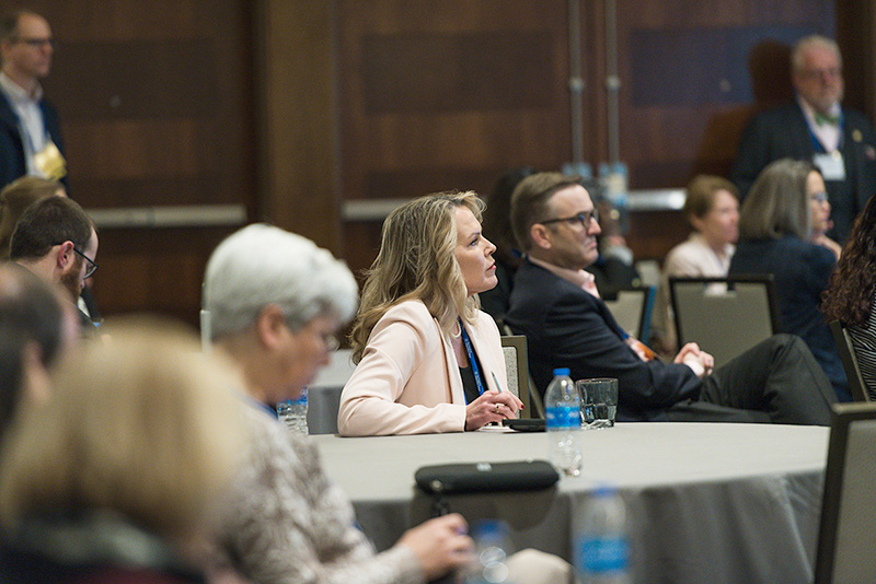 Participants seated at roundtables listen to speakers