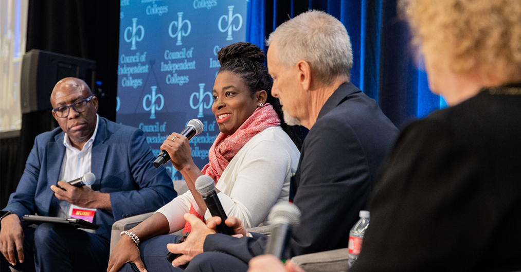 Four panelists holding microphones seated on stage present during plenary session